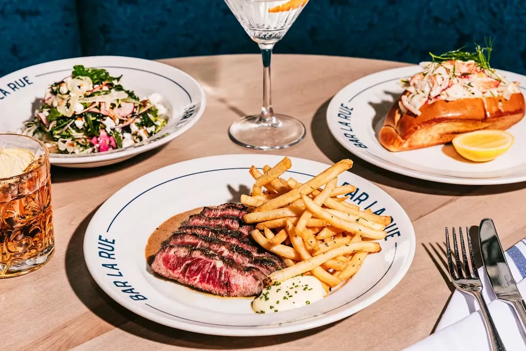 Steak frites, lobster roll, salad, and drinks on a wooden table.