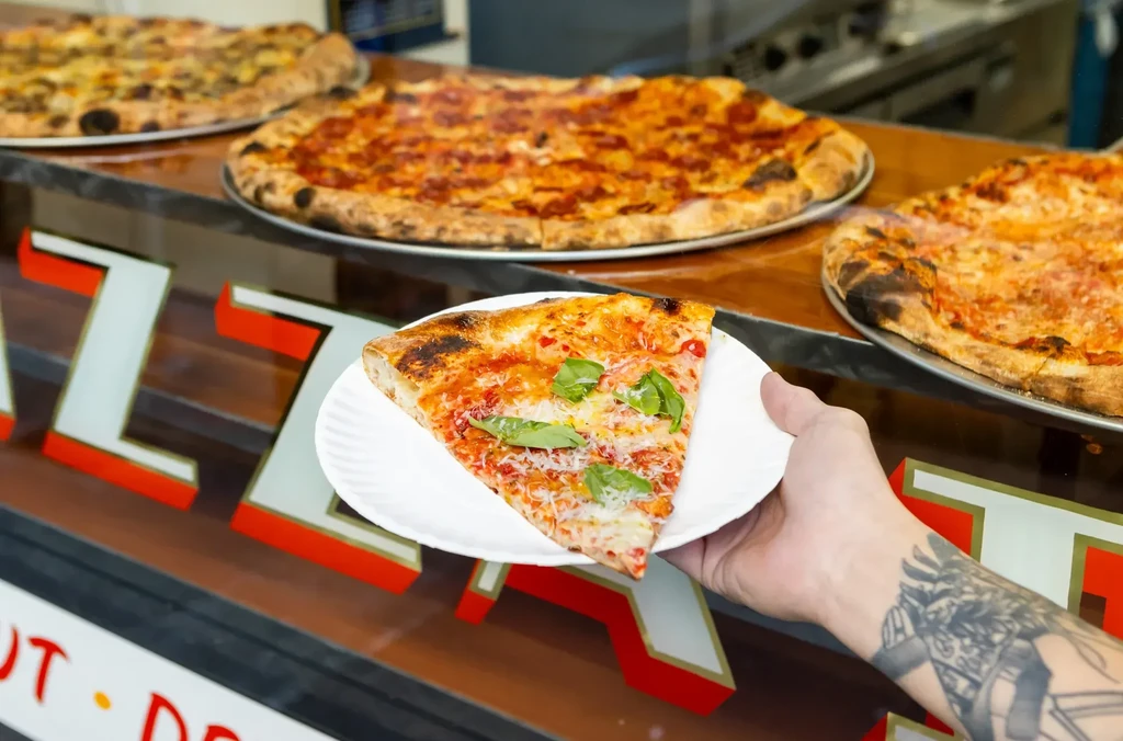A person with a tattooed arm holding a slice of pizza.