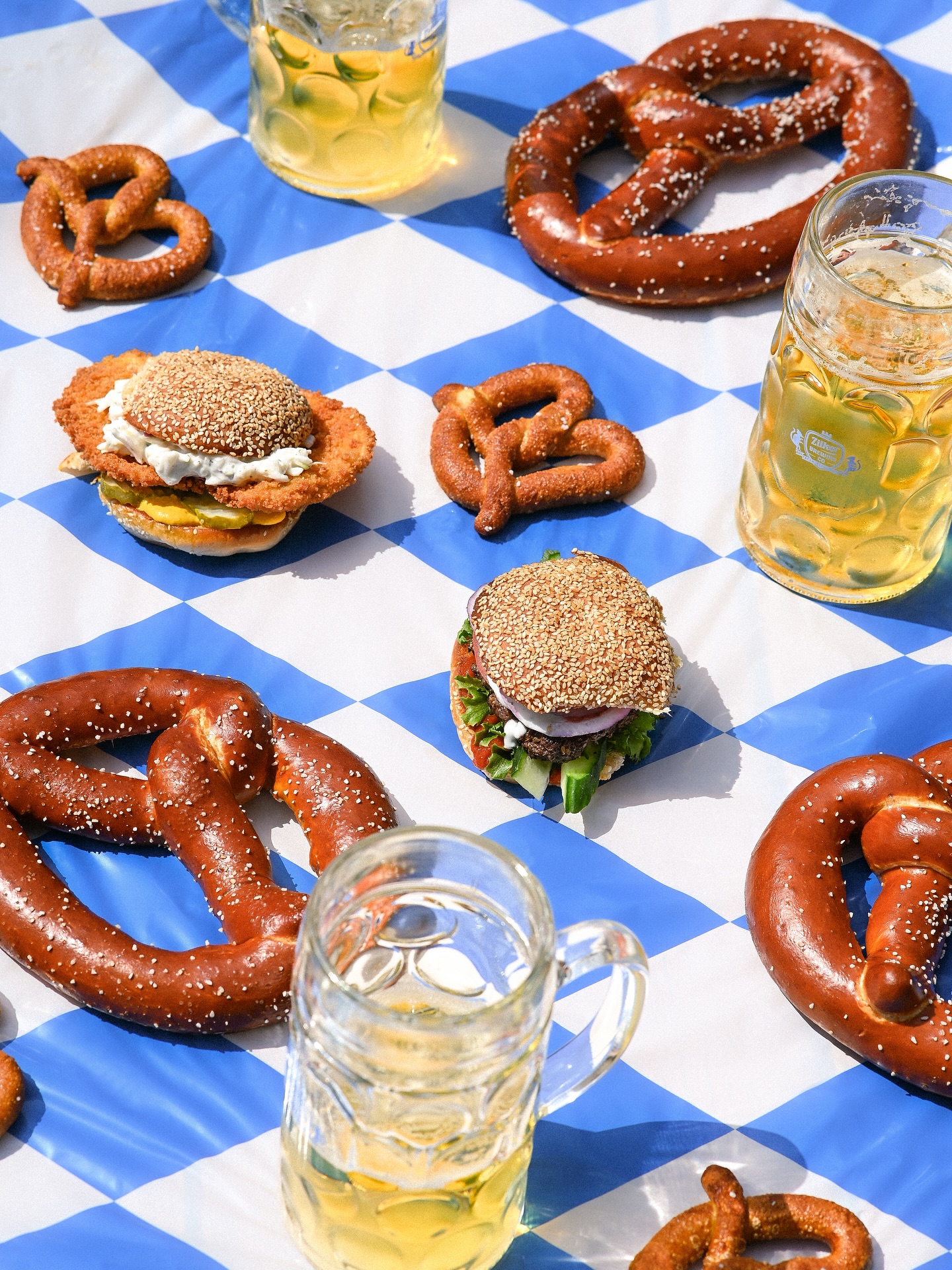 Pretzels, sandwiches, and beer mugs on blue and white checkered tablecloth.