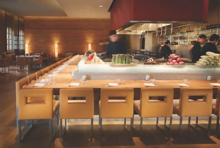 Long communal table in a restaurant with an open kitchen and chefs.