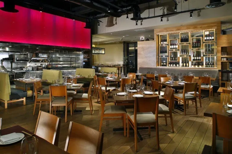 An empty restaurant with wooden tables and chairs, a kitchen, and a wine wall.