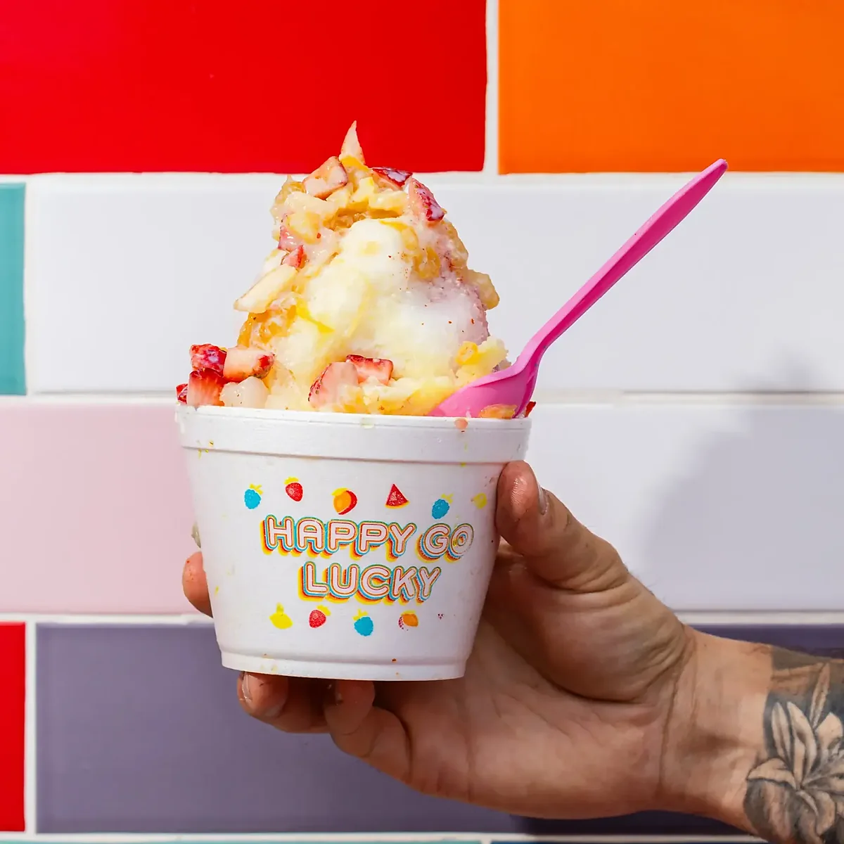 Hand holding cup of shave ice with strawberries against colorful tile wall