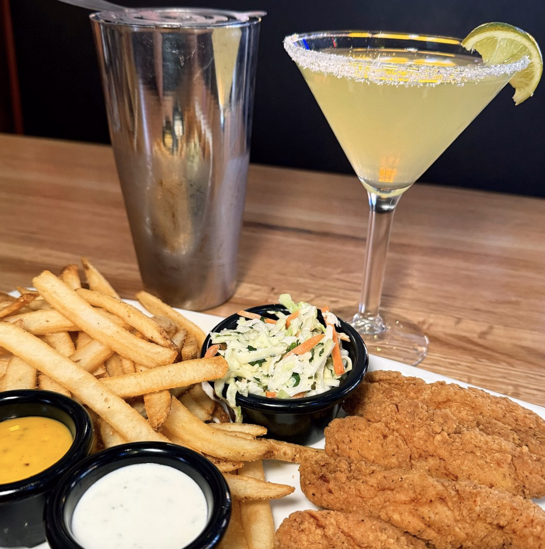Classic chicken tenders meal with fries, coleslaw, sauces, and a martini garnished with lime.