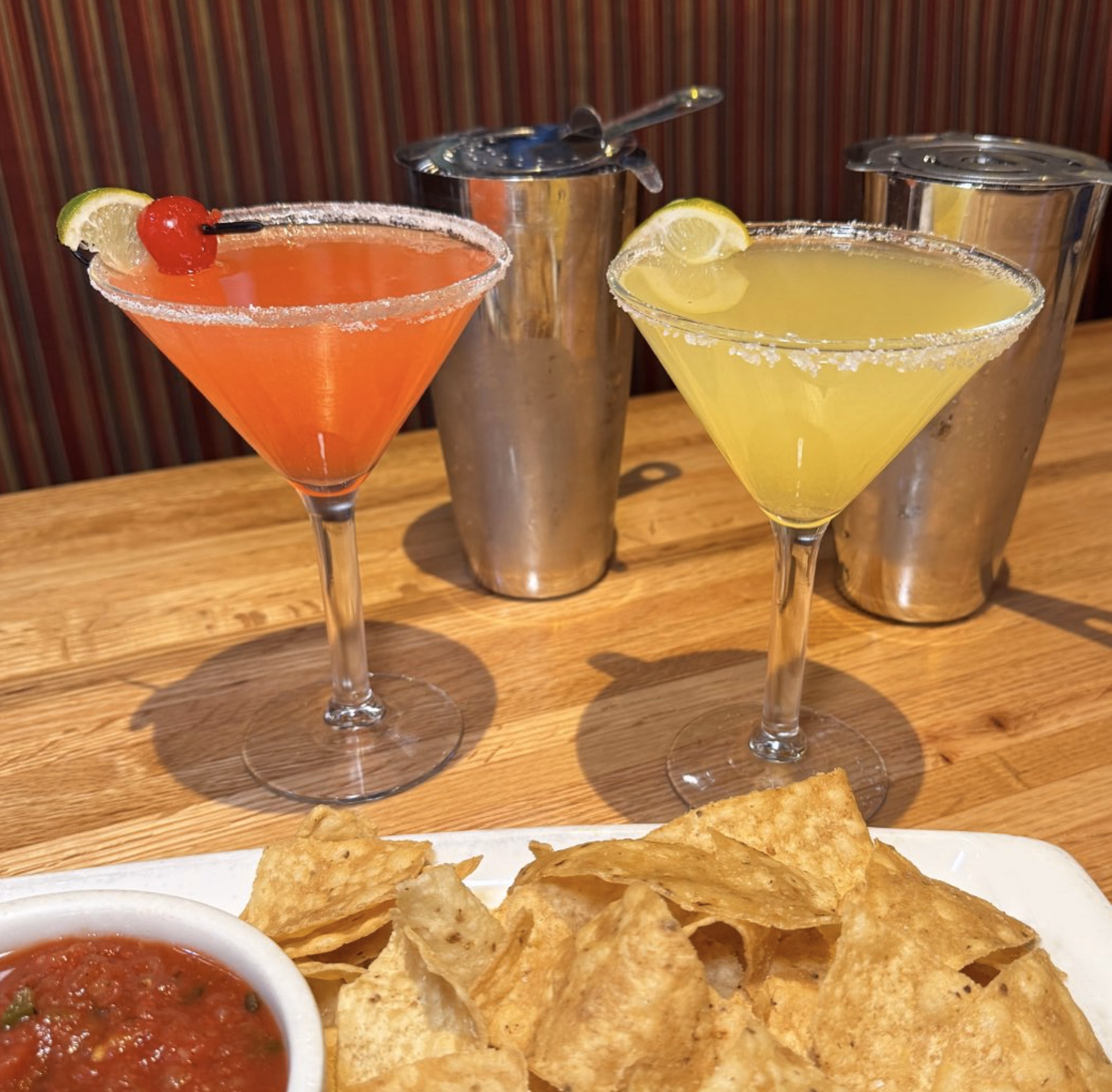 Two colorful cocktails with salted rims beside a basket of chips and fresh salsa.