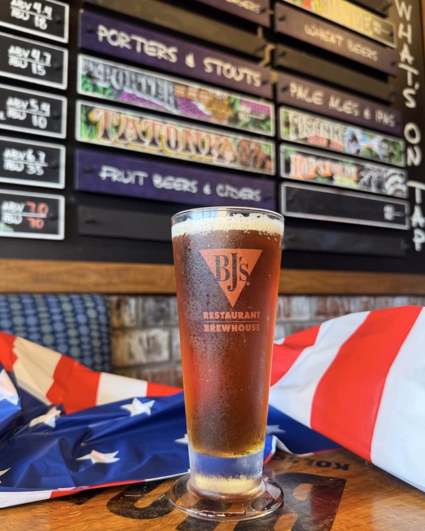 A tall glass of BJ’s craft beer placed in front of a colorful tap list of house brews.
