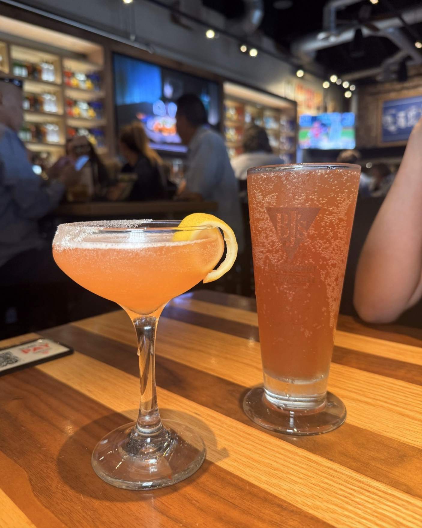 Two handcrafted cocktails on a wooden table at BJ’s Brewhouse with a lively bar scene in the background.