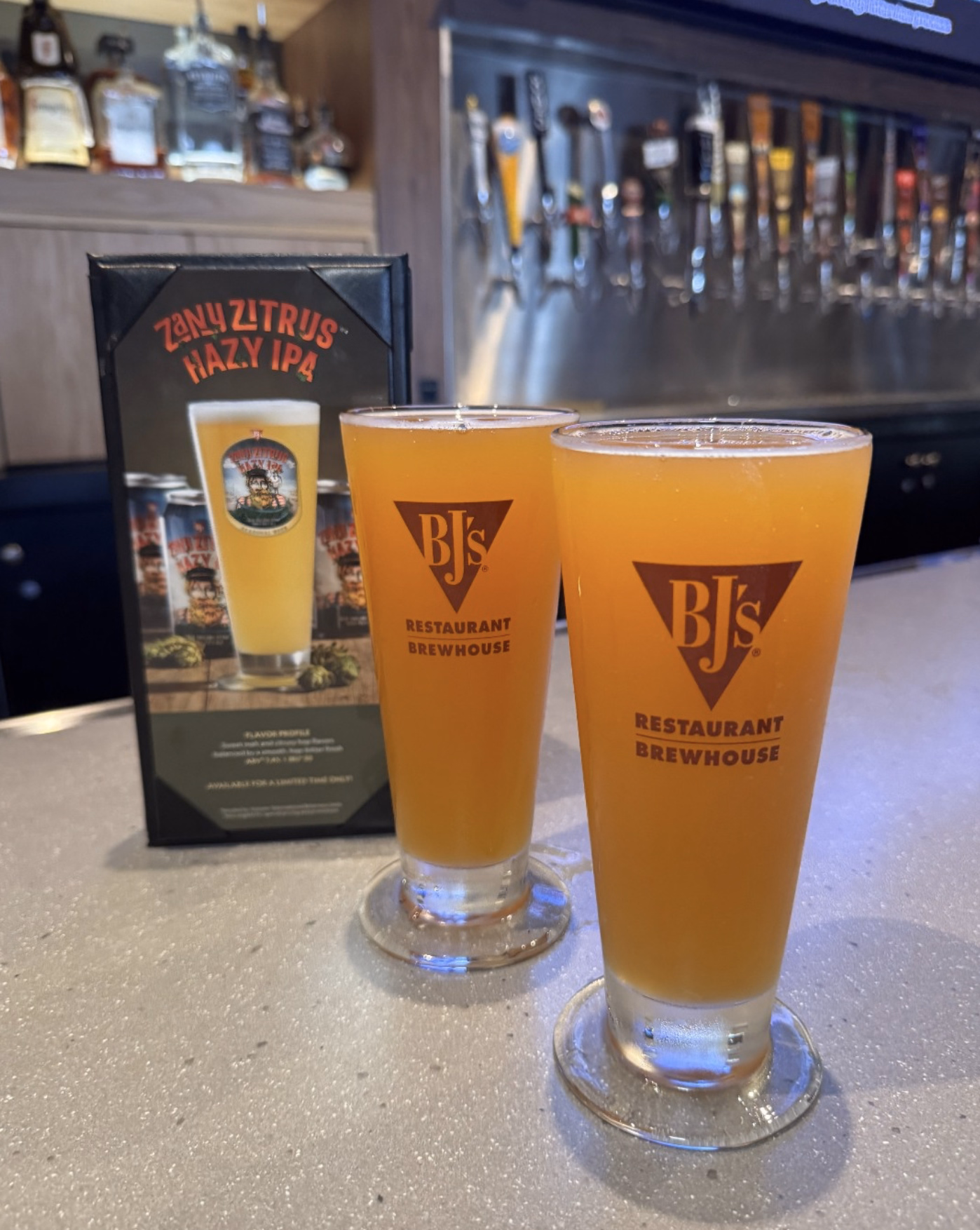 Two frosty glasses of BJ’s Zany Zitrus Hazy IPA on the bar with beer taps in the background.