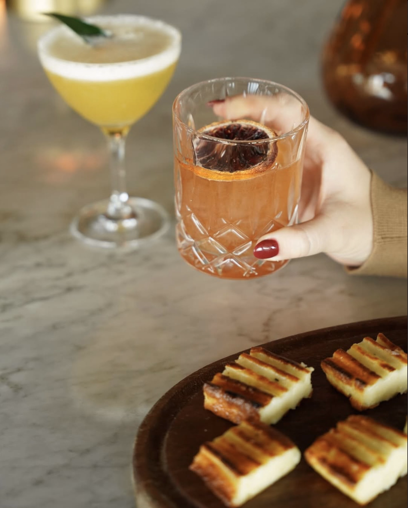 A hand holding a smoked citrus cocktail beside a passion fruit martini, with grilled cheese bites on a wooden board.