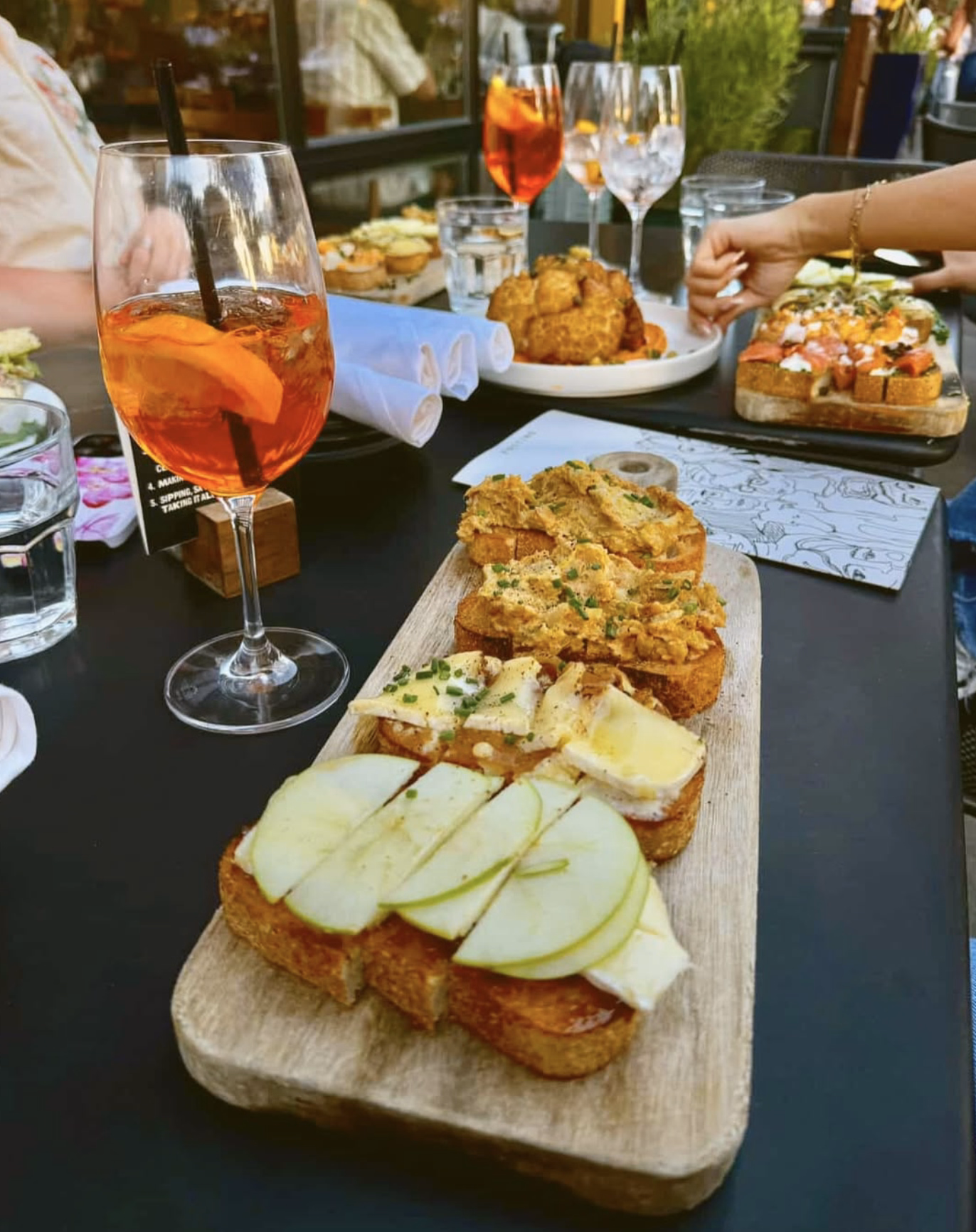 A close-up of gourmet bruschetta with fresh apple slices and cheese, surrounded by drinks and other shared plates on an outdoor dining setup.