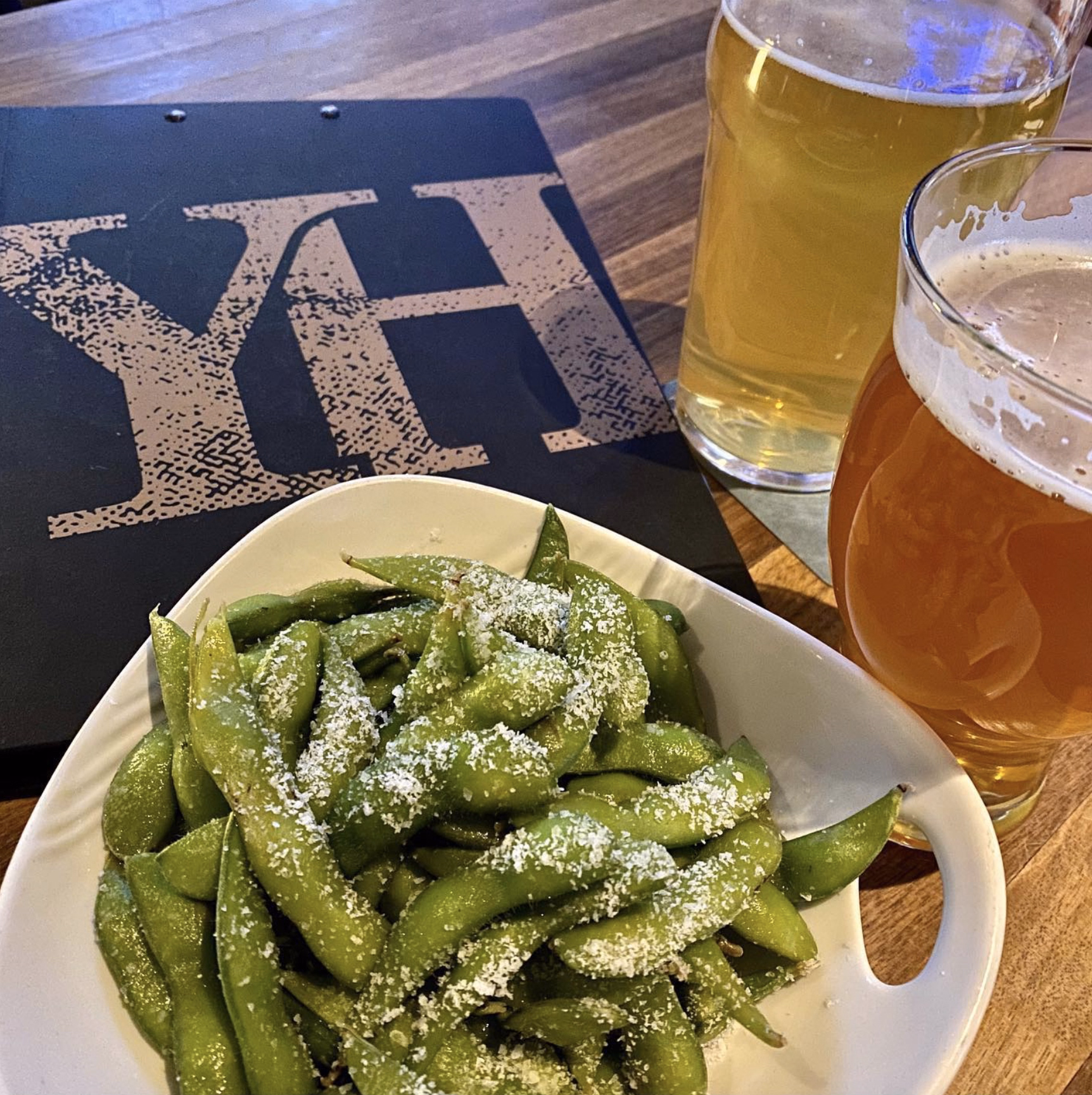 A bowl of salted edamame pods with two pints of beer on a wooden table.