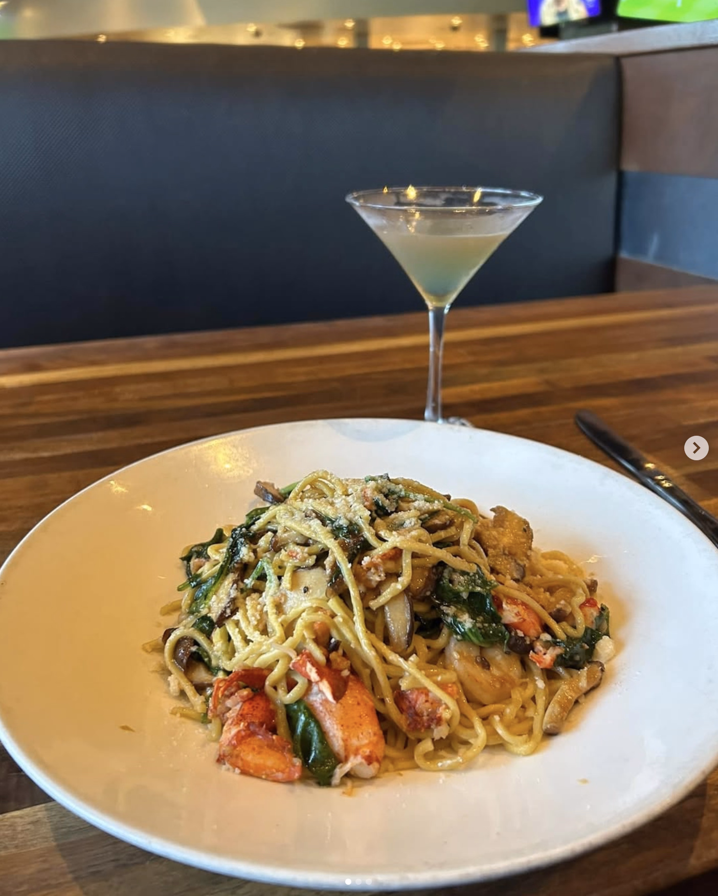 A plate of lobster pasta with spinach and mushrooms, topped with Parmesan cheese, paired with a martini on a wooden table.